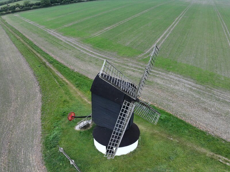 Aerial view of a traditional black weatherboarded post mill in open arable farmland, sails and external ladder visible
