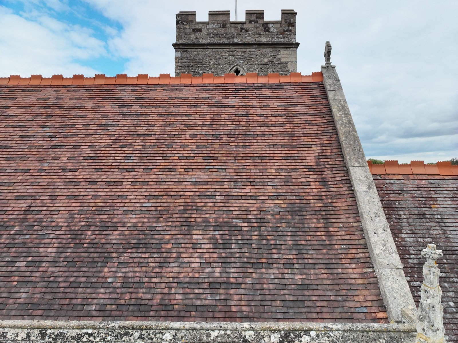 Oblique drone photograph across the tiled nave roof of All Saints church, Milton Keynes Village, with the battlemented stone tower beyond
