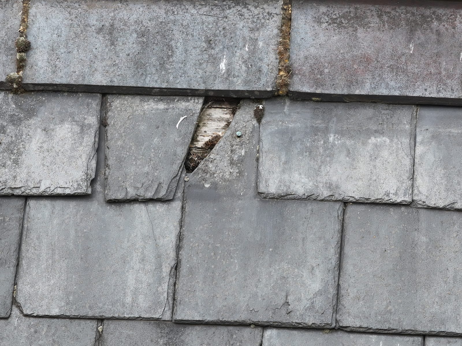 Close-range drone photograph of a slate roof slope at Christ Church, Chesham, where one slate has a large section broken away with timber boarding visible through the opening