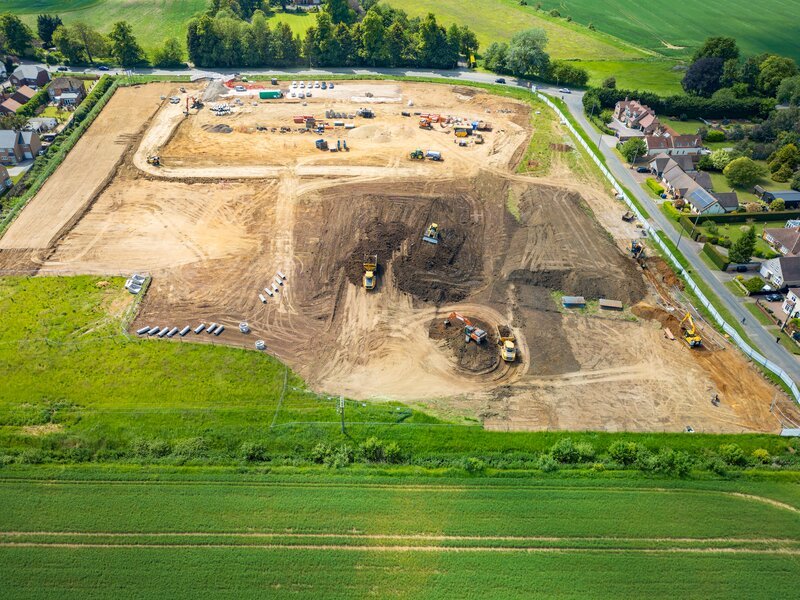 Aerial view of a UK construction site in groundworks phase, with excavators and dumpers cutting and filling, material stockpiles, drainage pipework laid out and access routes formed