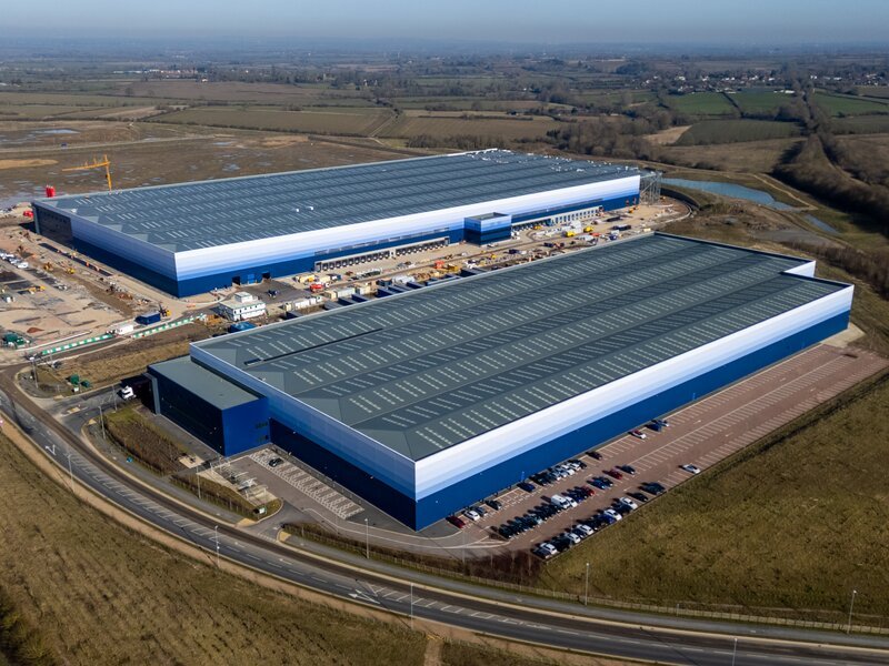 Aerial view of a large UK distribution and logistics complex with two blue-clad warehouses, loading docks and adjacent car parking