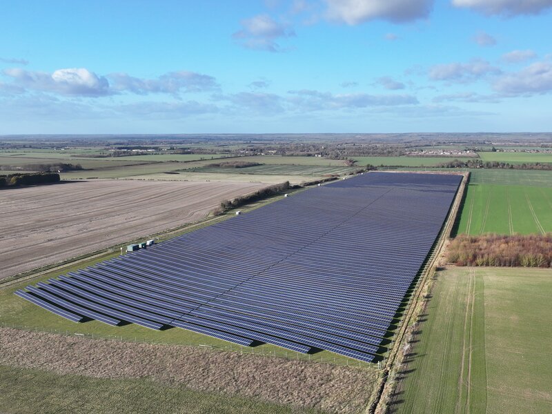 Aerial view of a UK solar farm with rows of photovoltaic panels in a fenced field, arable land and distant farmland visible beyond