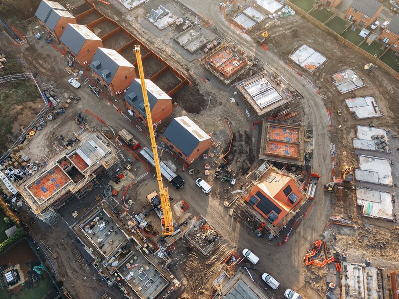 Top-down aerial view of a UK residential development under construction, with completed timber-frame homes, partial-build plots and a telehandler crane working between phases