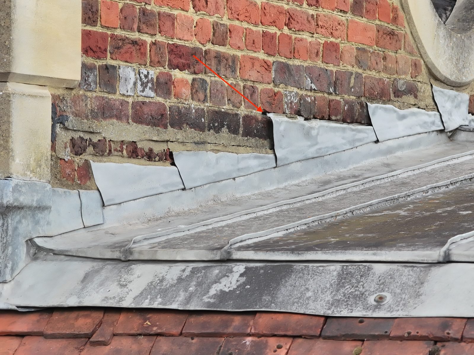 Close-range drone photograph of a lead parapet gutter at a church wall where the upper edges of the stepped lead flashings stand clear of the brick face, one position marked with a red arrow