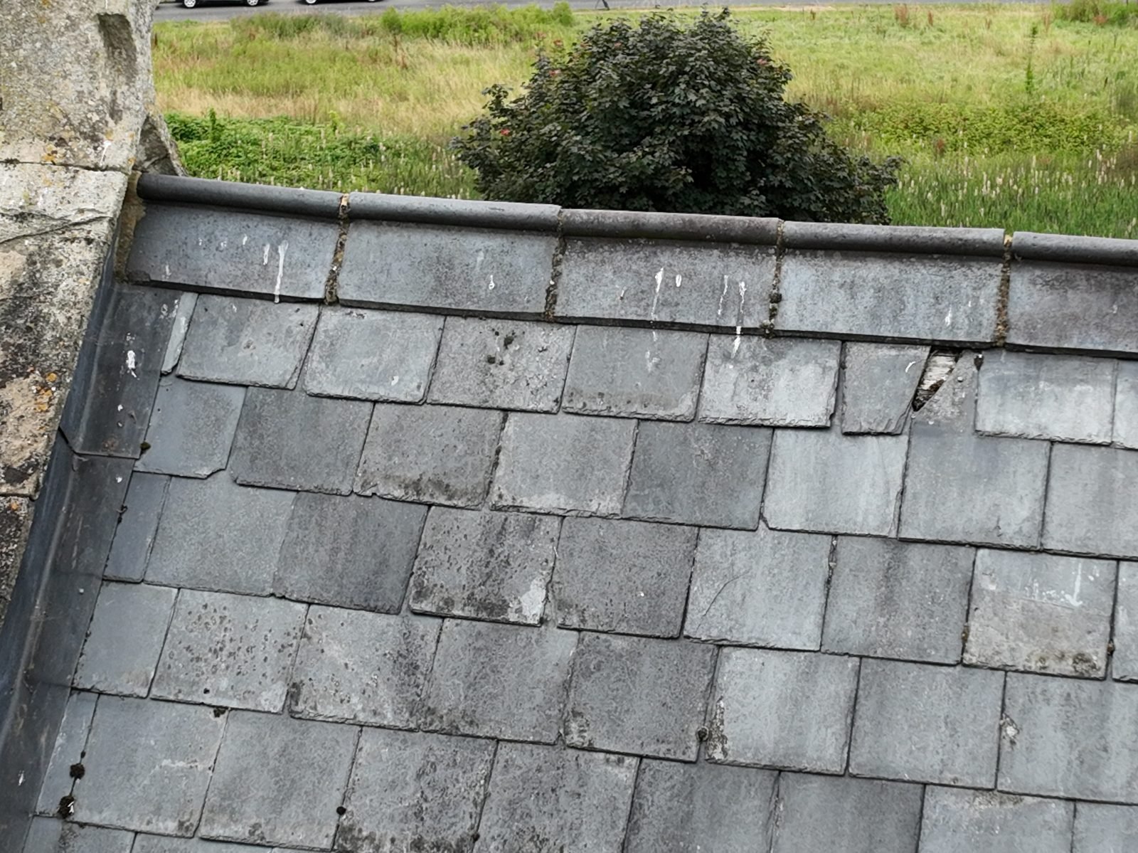 Close-range drone photograph of the head of a slate roof slope beside a stone gable, showing the ridge capping, a slipped slate at the top course and the churchyard beyond